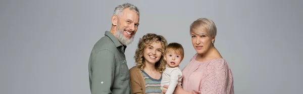 Joyful middle aged couple smiling near adult daughter with toddler girl isolated on grey, banner — Stock Photo