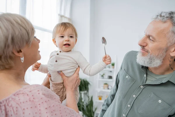 Woman holding cheerful granddaughter with spoon near bearded grey haired husband in kitchen — Stock Photo