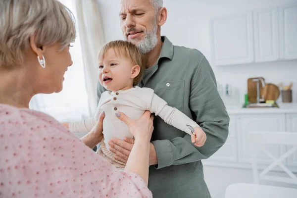 Bearded man holding toddler granddaughter near blurred wife in kitchen — Stock Photo