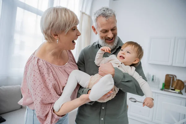 Cheerful middle aged couple having fun with smiling granddaughter holding spoon in kitchen — Stock Photo