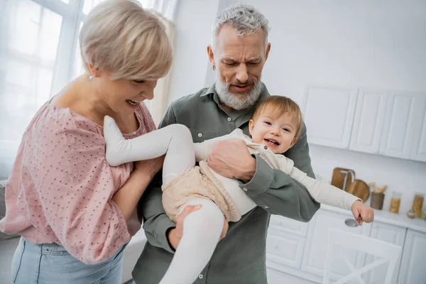Carefree baby girl holding spoon near smiling grandparents in kitchen — Stock Photo