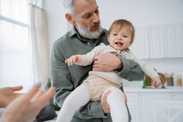 Mature bearded man holding cheerful toddler granddaughter in kitchen — Stock Photo