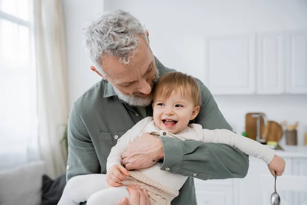 Bearded mature man embracing carefree granddaughter holding spoon in kitchen — Stock Photo