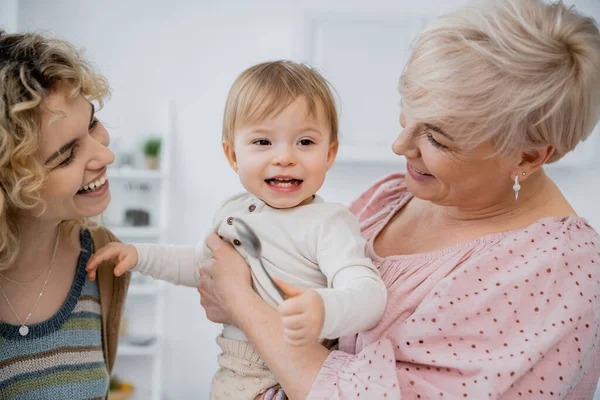 Carefree baby girl holding spoon near cheerful mom and granny in kitchen — Stock Photo