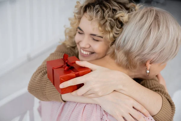 Überglückliche Frau mit geschlossenen Augen hält Geschenkbox und umarmt Mutter mittleren Alters zu Hause — Stockfoto