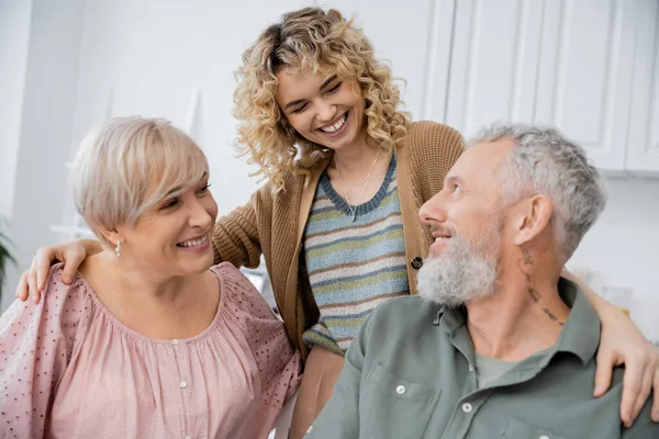 Smiling woman with blonde wavy hair embracing joyful middle aged parents in kitchen at home — Stock Photo