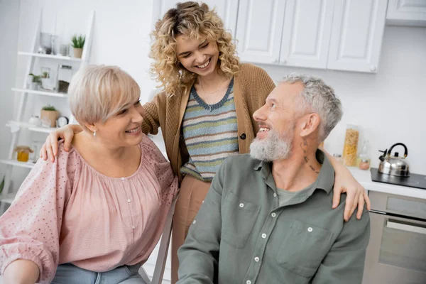 Cheerful woman with curly blonde hair hugging happy parents in kitchen — Stock Photo