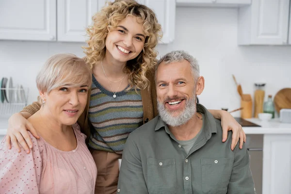 Complacida pareja de mediana edad sonriendo a la cámara cerca de hija adulta en la cocina en casa - foto de stock