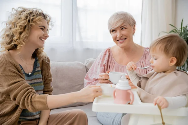 Happy mature woman with tea cup looking at adult daughter feeding toddler girl in kitchen — Stock Photo