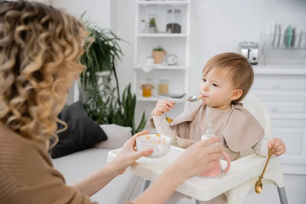 Blurred woman holding bowl and baby bottle near toddler daughter eating breakfast in kitchen — Stock Photo