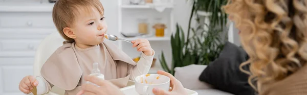 Little child with spoon eating puree near blurred mother holding bowl in kitchen, banner — Stock Photo