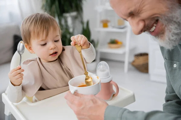 Blurred man laughing near little granddaughter holding spoons near bowl with puree in kitchen — Stock Photo