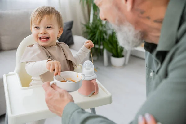 Carefree girl with spoons sitting on baby chair near bowl with breakfast and grandpa on blurred foreground in kitchen — Stock Photo