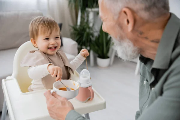 Cheerful baby holding spoon near bowl with puree and bearded grandfather on blurred foreground in kitchen — Stock Photo