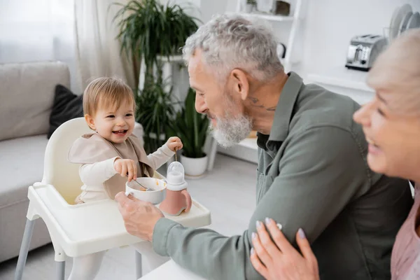 Pareja madura sonriendo cerca despreocupada nieta con cucharas sentado en silla de bebé cerca de tazón con desayuno en la cocina - foto de stock