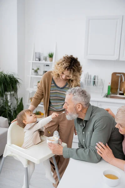 Toddler girl with spoon feeding bearded grandfather near smiling family in kitchen — Stock Photo