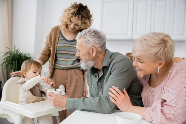 Baby girl holding spoon near bowl while having breakfast near mother and happy grandparents in kitchen — Stock Photo