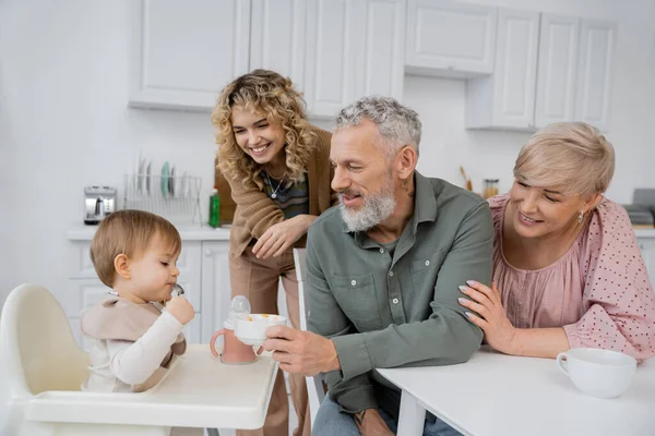 Усміхнений бородатий чоловік тримає миску біля онуки, сидячи на дитячому стільці під час сніданку на кухні — стокове фото