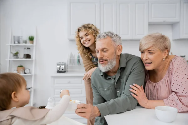 Smiling bearded man holding bowl while feeding kid with breakfast near family in kitchen — Stock Photo