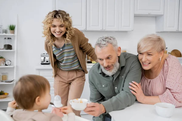 Веселий бородатий чоловік тримає миску біля розмитої онуки і усміхненої сім'ї на кухні — стокове фото