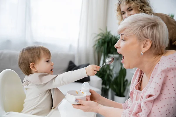 Side view of little child with spoon feeding grandmother with bowl in hands — Stock Photo