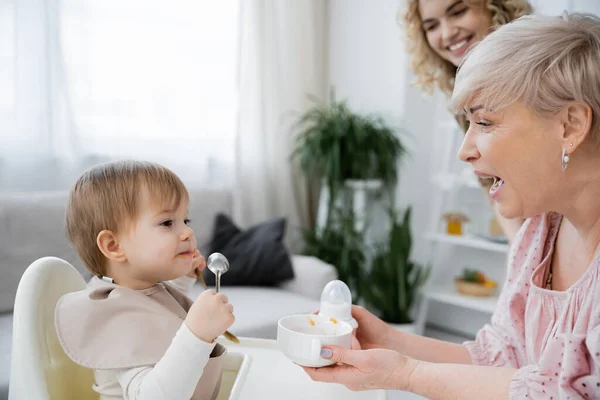 Reife Frau öffnet Mund, während sie Schüssel in der Nähe Enkelin mit Löffel beim Frühstück in der Küche hält — Stockfoto