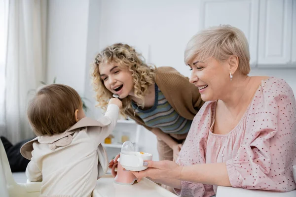 Toddler girl feeding mom near smiling grandmother with bowl in kitchen — Stock Photo