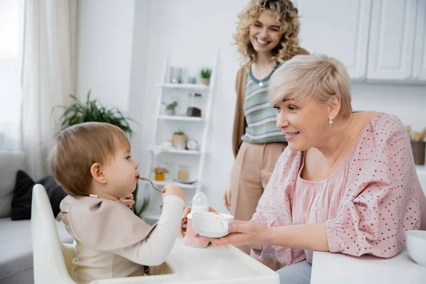 Pleased middle aged woman holding bowl near toddler granddaughter with spoon during breakfast in kitchen — Stock Photo