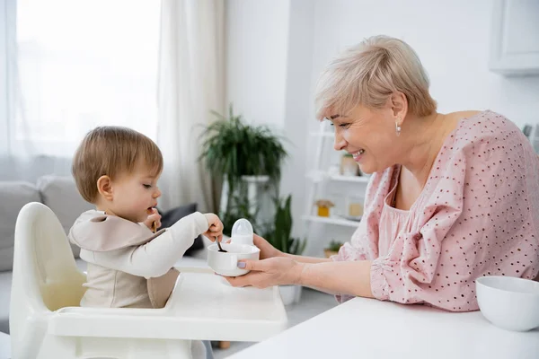 Side view of smiling middle aged woman holding bowl near granddaughter sitting on baby chair and having breakfast in kitchen — Stock Photo