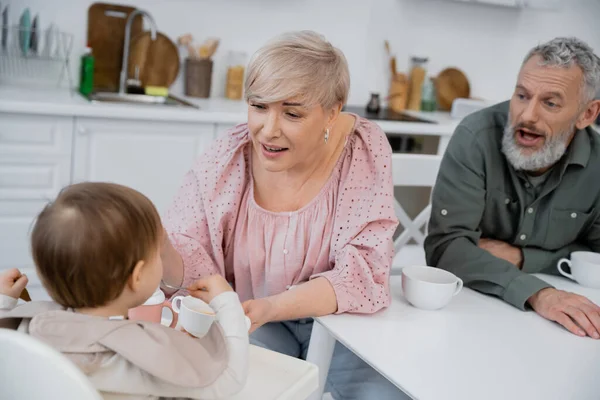 Happy woman holding bowl near granddaughter eating breakfast in kitchen while bearded man opening mouth on blurred background — Stock Photo