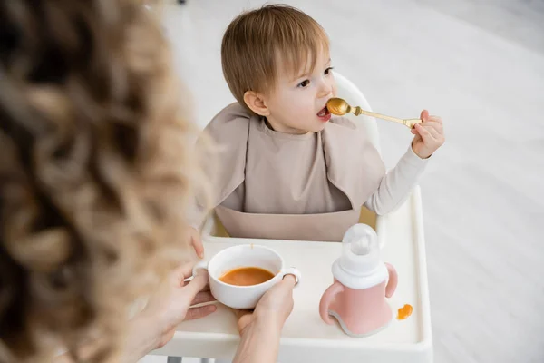 Bambino ragazza seduta sulla sedia del bambino e mangiare purea vicino a mamma offuscata con ciotola in cucina — Foto stock