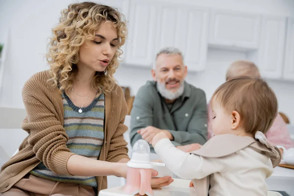 Donna che nutre piccola figlia con colazione vicino ai genitori maturi sorridendo su sfondo sfocato in cucina — Foto stock