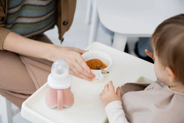 Vista cortada da mulher segurando tigela com purê perto da filha da criança sentada na cadeira do bebê durante o café da manhã — Fotografia de Stock
