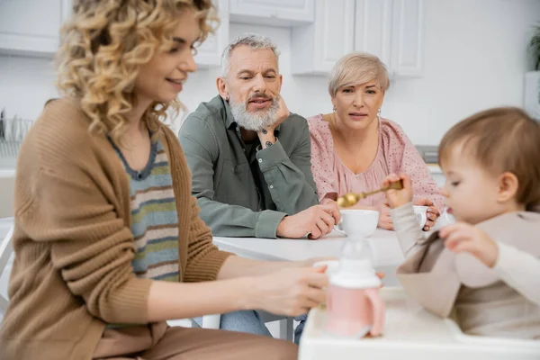 Middle aged couple looking at happy daughter feeding toddler girl with breakfast in kitchen — Stock Photo