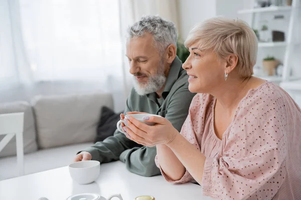 Pleased middle aged woman with cup of tea looking away near smiling bearded husband in kitchen — Stock Photo