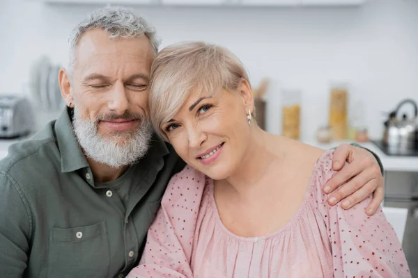 Hombre barbudo feliz con los ojos cerrados abrazando esposa sonriendo a la cámara en la cocina - foto de stock