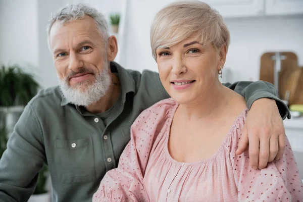 Heureuse femme d'âge moyen et homme barbu souriant à la caméra tout en étant assis dans la cuisine à la maison — Photo de stock