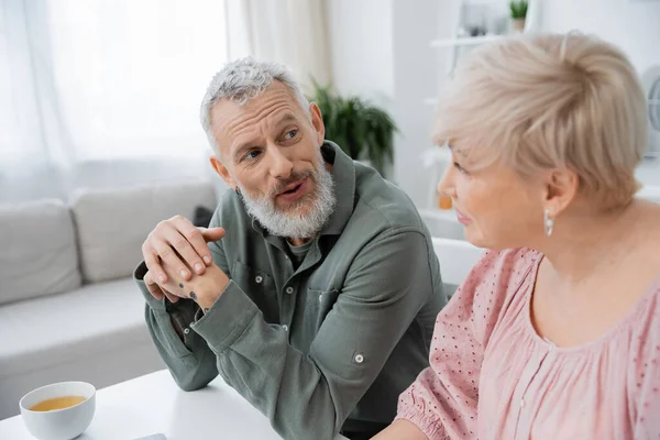 Позитивний бородатий чоловік розмовляє з розмитою дружиною середнього віку біля чашки чаю на столі на кухні — стокове фото