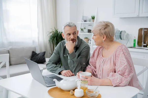 Barbudo hombre de mediana edad mirando a la esposa mientras habla cerca de taza de té y portátil en la mesa en la cocina moderna - foto de stock