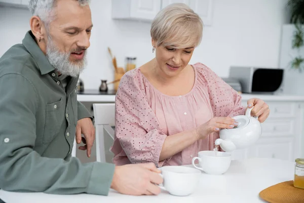Alegre mujer vertiendo té cerca barbudo marido sonriendo en la cocina - foto de stock