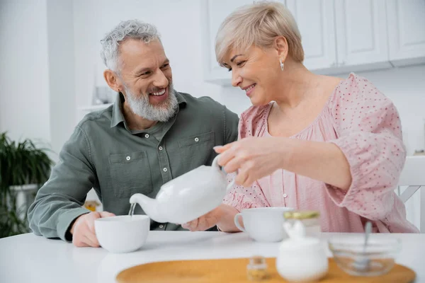 Glückliche Frau mittleren Alters gießt Tee neben fröhlichem bärtigen Ehemann in der Küche — Stockfoto