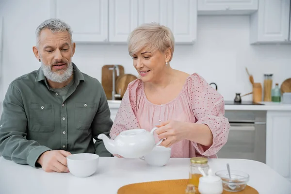 Happy middle aged woman pouring tea near bearded husband during breakfast in kitchen — Stock Photo