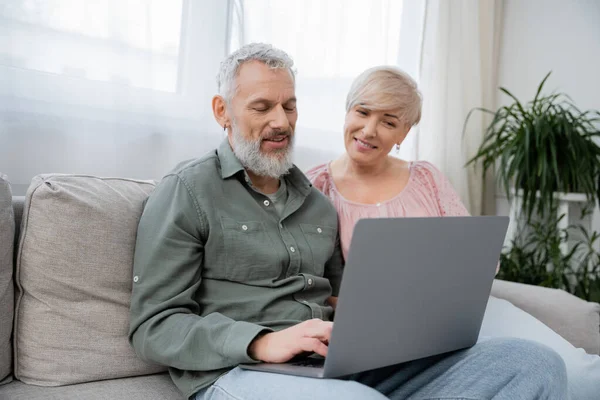 Позитивная зрелая пара смотрит фильм на ноутбуке, сидя на диване в гостиной — стоковое фото