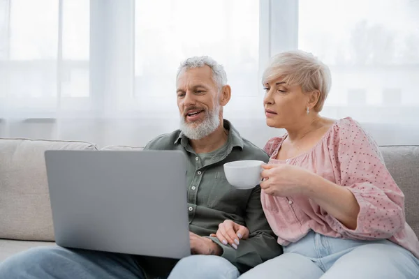 Mulher de meia idade segurando xícara de chá enquanto assiste filme no laptop perto do marido sorridente no sofá na sala de estar — Fotografia de Stock