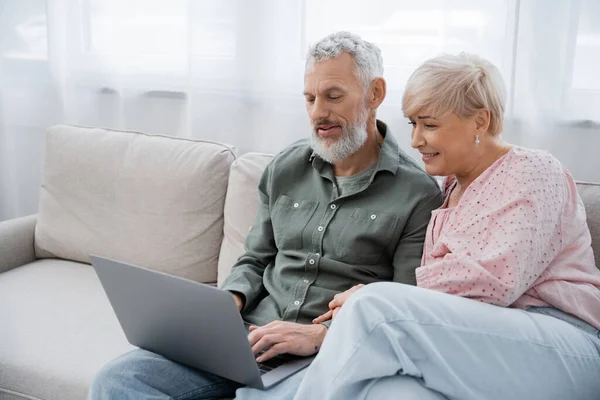 Радісна жінка середнього віку і бородатий чоловік сидить на дивані у вітальні і дивиться фільм на ноутбуці — стокове фото