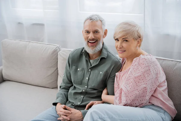 Middle aged woman with excited bearded husband sitting on sofa in living room and smiling at camera — Stock Photo