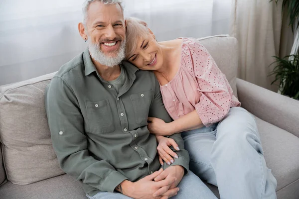 Feliz mulher de meia-idade apoiando-se no homem barbudo alegre sorrindo para a câmera no sofá na sala de estar — Fotografia de Stock