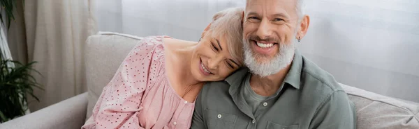 Spensierato uomo di mezza età che sorride alla telecamera vicino alla moglie felice sul divano in soggiorno, banner — Foto stock