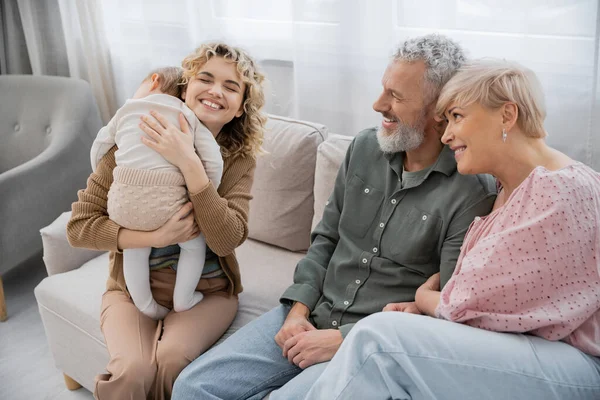 Overjoyed woman with closed eyes embracing toddler daughter while spending time with parents on couch at home — Stock Photo