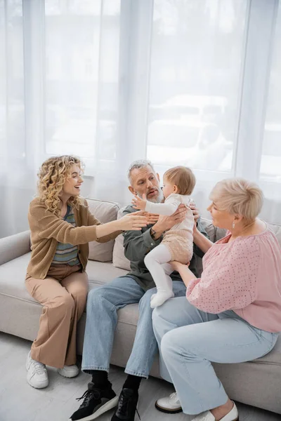 Bearded man holding toddler granddaughter near wife and happy daughter on sofa in living room — Stock Photo